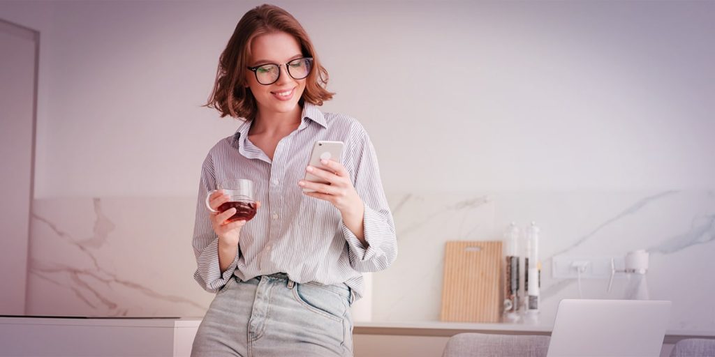 mulher tomando café encostada em mesa de escritório conferindo no celular como vai seu marketing digital